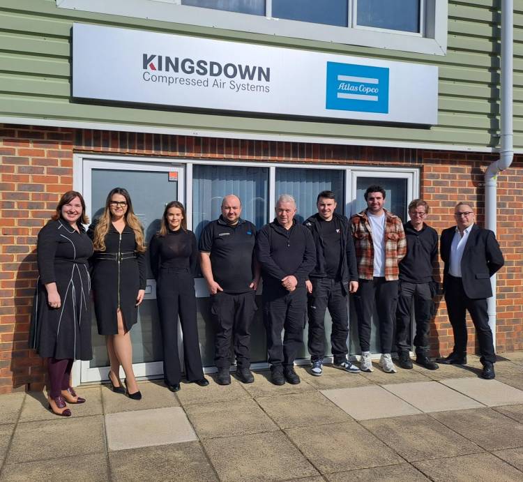 A group of nine individuals stand together outdoors in front of a building entrance, posed for a team photo. The background shows an office building with a green horizontal siding facade above a brick ground level. Above the glass doors and windows, a sign reads "KINGSDOWN Compressed Air Systems" in black text with a blue and white logo. To the right of the text on the sign is a separate blue and white "Atlas Copco" logo. The group is diverse in age and dress, with individuals in business attire and casual workwear standing close together on a tiled sidewalk, smiling towards the camera. The setting is bright and sunny, casting distinct shadows on the ground. The pavement in front of the group is composed of light-colored square paving slabs with darker gray square accents.
